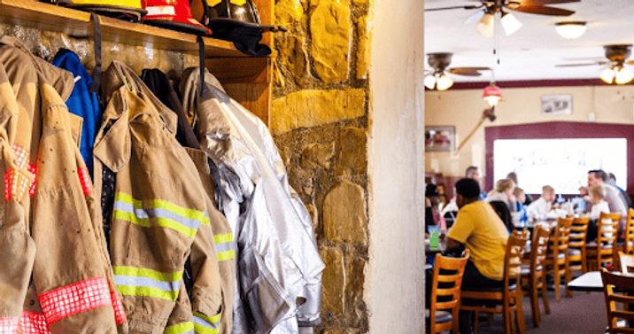 Fire fighter jackets and hats hanging at entrance of hook and ladder restaurant with people seated in background at tables